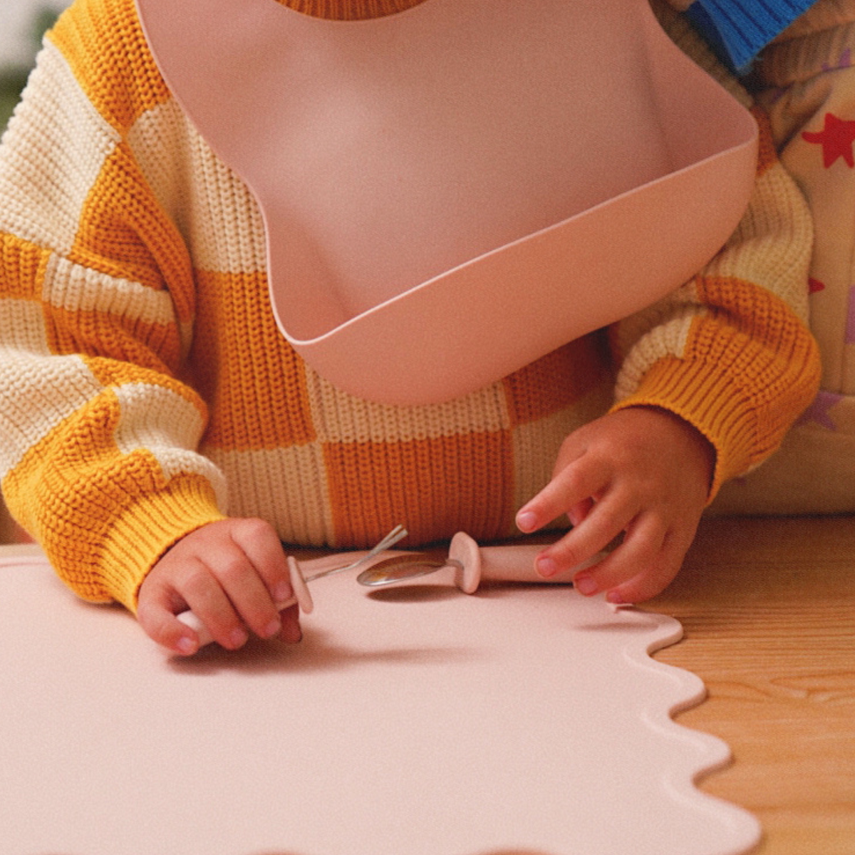 Child wearing a silicone pink baby bib designed by Rommer, playing with cutlery at the table