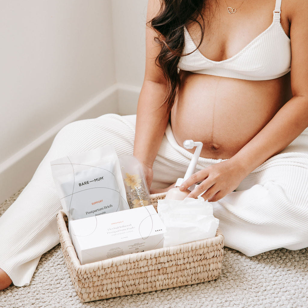 Pregnant woman sitting on the floor with a basket of Bare Mum Postpartum products.