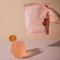 Hand holding a pink package of franjos kitchen blood orange hydration powder next to a glass of pink drink.