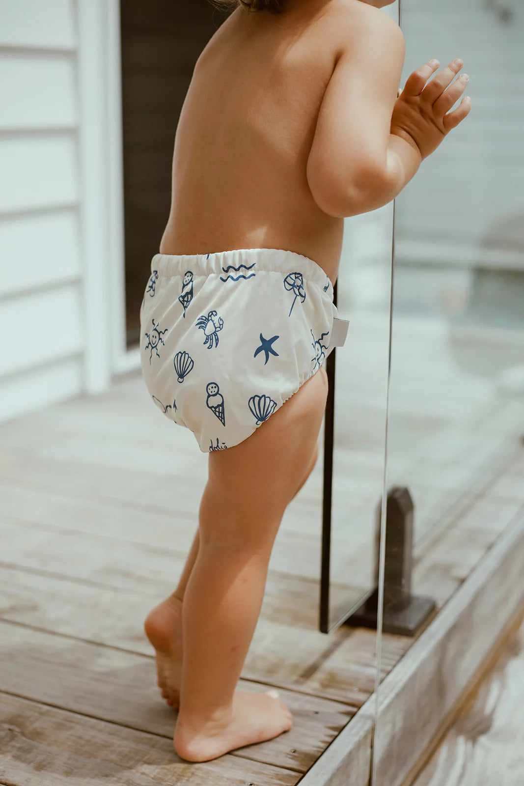 Child wearing a reusable swim nappy with sea-themed print on a wooden deck.