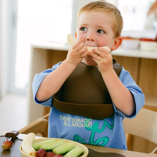 Child eating a banana while wearing a brown Rommer baby bib