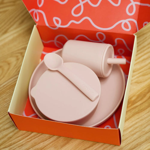 Pink Rommer children's dinner set including silicone plate, bowl, and spoon in an open red box with white patterns on a wooden surface.