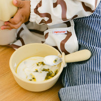Child eating yogurt with Rommer kids cutlery from a silicone bowl, wearing a striped apron.