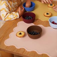 Children eating food on a wooden table using colorful silicone placemats and bowls designed by Rommer