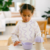 Child wearing a Rommer smock bib with a lilac wavy pattern in a home setting