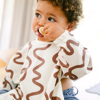 Child wearing a Rommer smock bib with brown wavy patterns, eating yogurt.