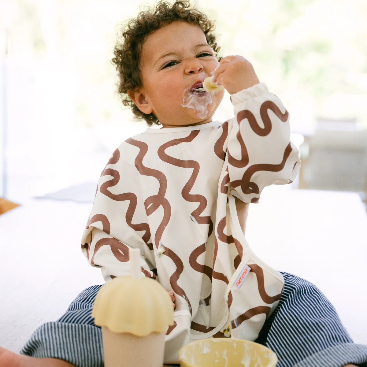 Child wearing a brown wavy patterned Smock bib eating a snack indoors.