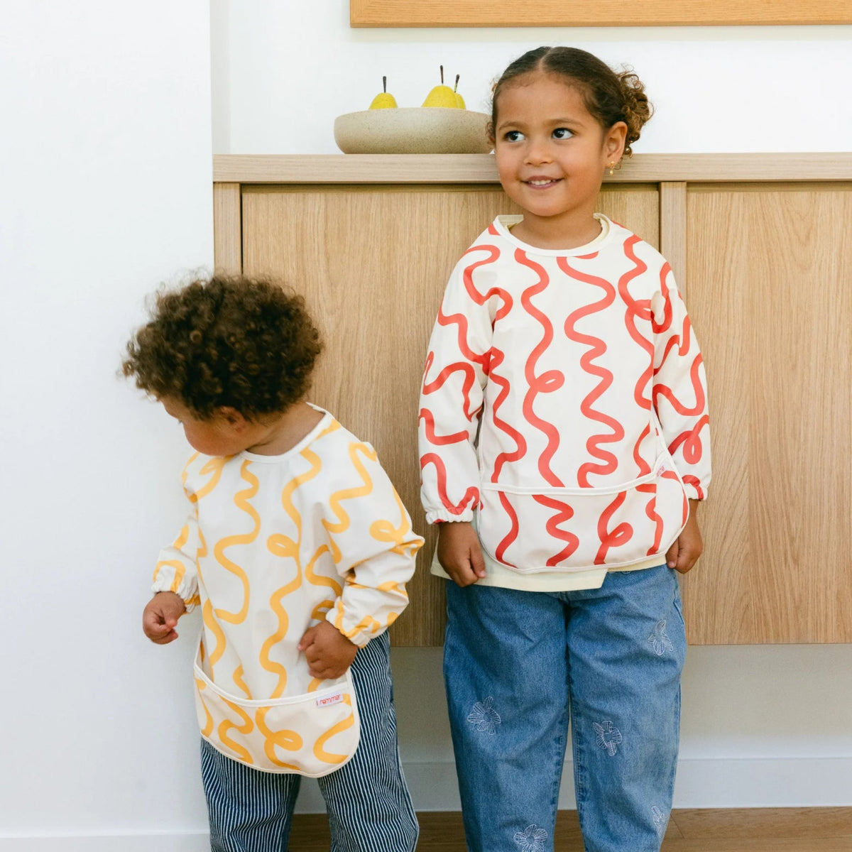 Two children wearing colourful smock bibs designed by Rommer standing in a kitchen.