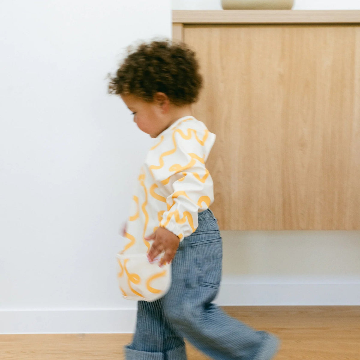 Child wearing a Rommer smock bib with yellow patterns and blue jeans walking indoors.