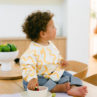 Child wearing a Rommer smock bib with yellow patterns sitting on a table eating from a silicone dinner set